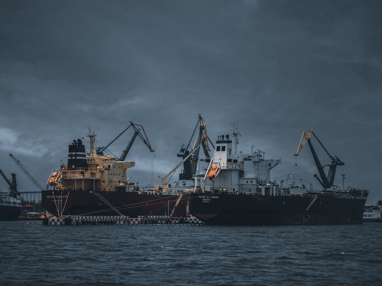Cargo ships at a bustling port under cloudy skies, cranes ready for loading.